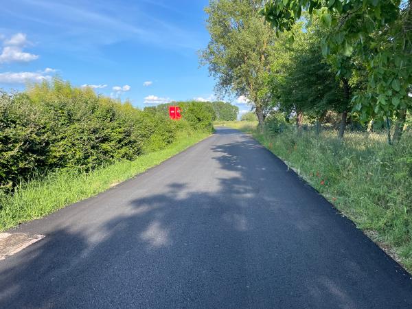 Paved bicycle path in rural area, hedge on left and trees with bushes on right bordering another road with visible vehicle.